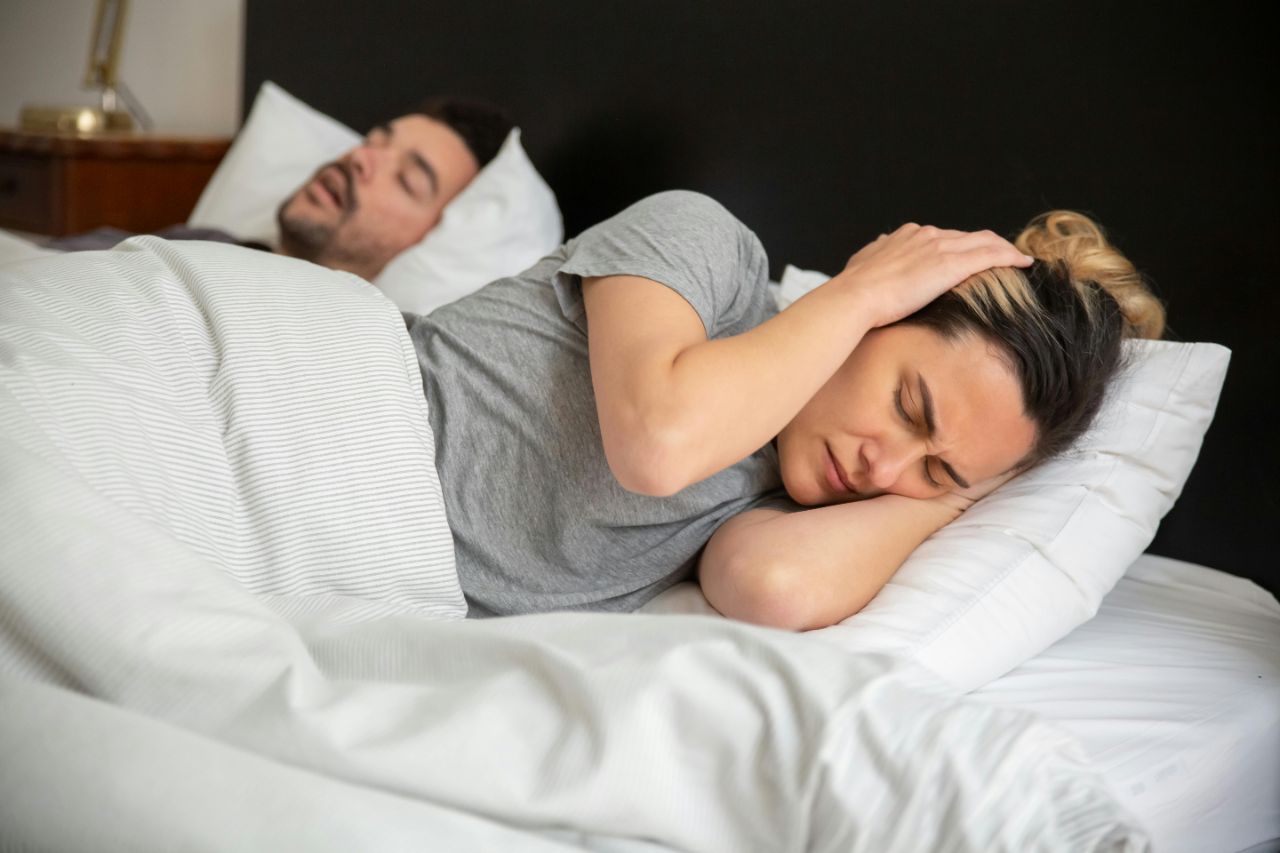 Ein Paar liegt in einem Bett mit weißer Bettwäsche, die Frau hält sich mit den Händen die Ohren zu während der Mann schläft.