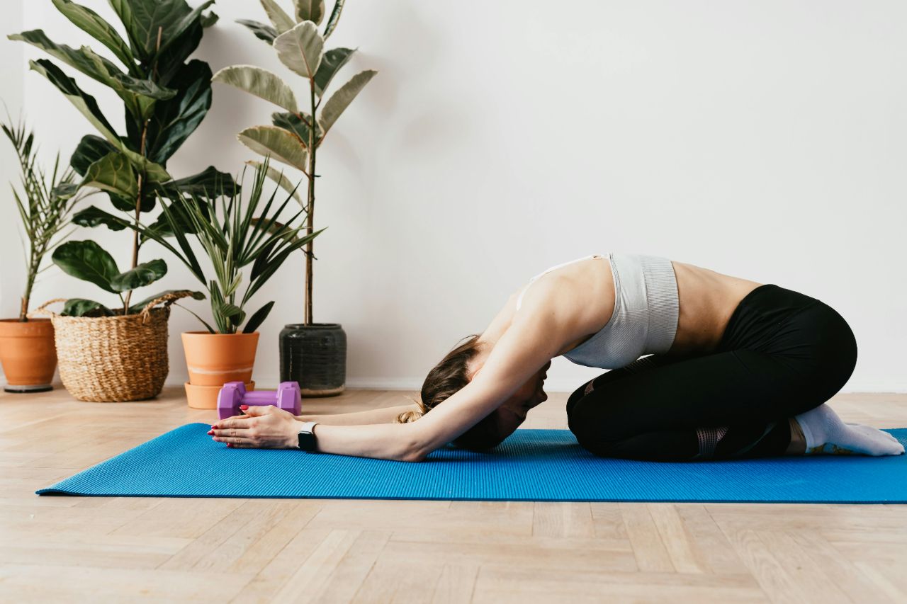 Frau in Sportbekleidung macht eine Übung auf einer Yogamatte, neben ihr liegen Hanteln und im Hintergrund stehen Pflanzen.