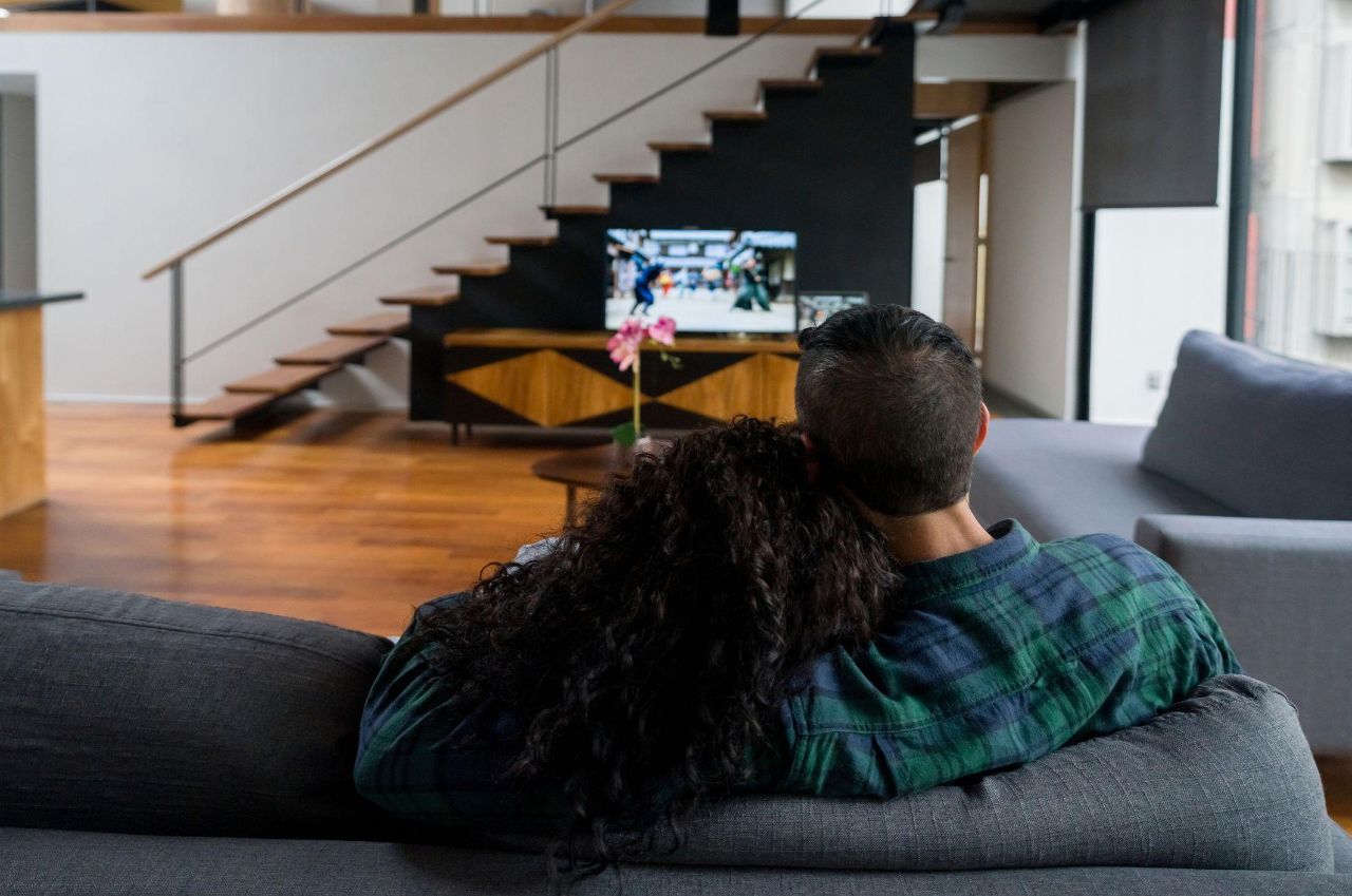 Ein von hinten abgebildetes Paar sitzt zusammen auf einer anthrazitfarbenen Couch im Wohnzimmer und schaut Fernsehen