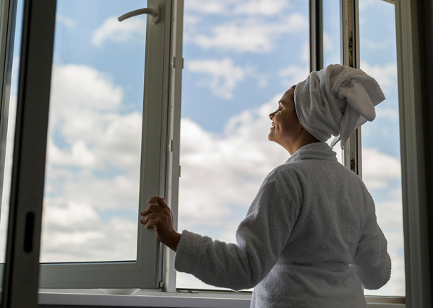 Frau im weißen Bademantel und Haarturban steht lächelnd vor dem weit geöffneten Fenster und genießt die frische Luft